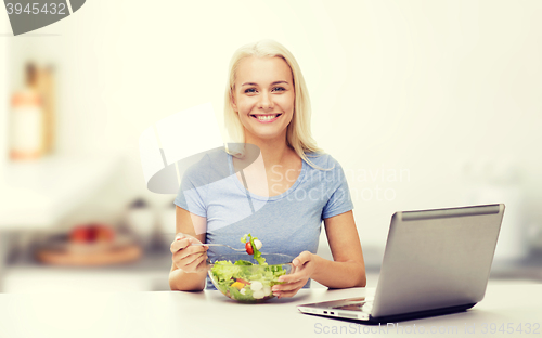 Image of smiling woman eating salad with laptop on kitchen