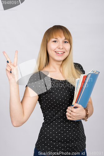 Image of The joyful girl with a folder in his hands shows gesture \"Victoria\" victory hand