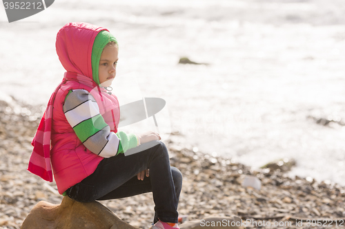 Image of Thoughtful girl sitting on a rock on the stony beach of the sea and looks into the distance