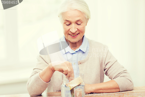 Image of senior woman putting money into glass jar at home