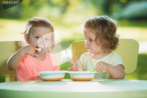 Image of Two little girls sitting at a table and eating together against green lawn