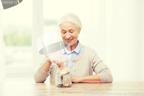 Image of senior woman putting money into glass jar at home