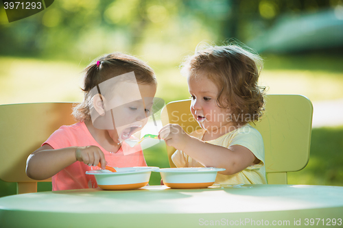 Image of Two little girls sitting at a table and eating together against green lawn