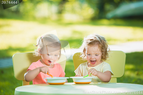 Image of Two little girls sitting at a table and eating together against green lawn