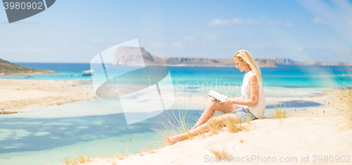 Image of Woman reading book, enjoying sun on beach.