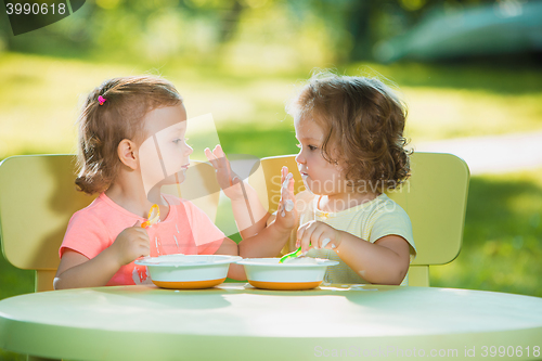 Image of Two little girls sitting at a table and eating together against green lawn