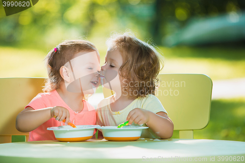 Image of Two little girls sitting at a table and eating together against green lawn