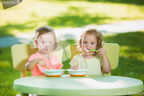 Image of Two little girls sitting at a table and eating together against green lawn