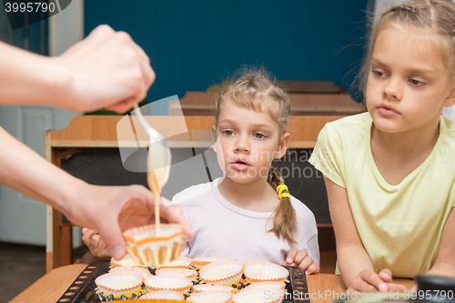 Image of Two girls with a surprised look like mom pours batter into molds for cupcakes