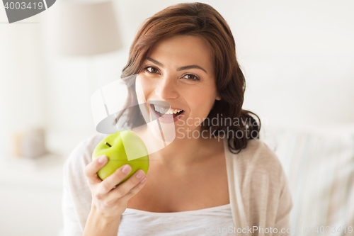 Image of smiling young woman eating green apple at home