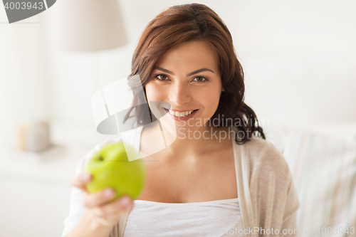 Image of smiling young woman eating green apple at home