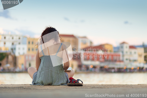 Image of Rear view of woman sitting on a pier, reading book.