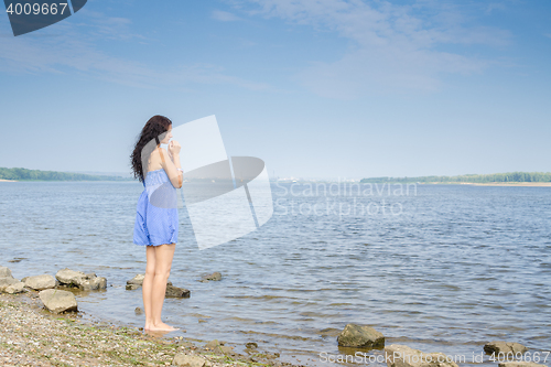 Image of Sad young brunette woman in a blue summer dress standing on the river bank