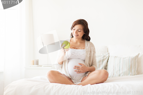 Image of happy pregnant woman eating green apple at home