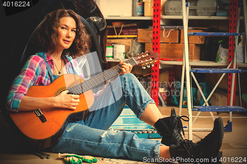 Image of beautiful Woman mechanic in blue overalls resting with a guitar