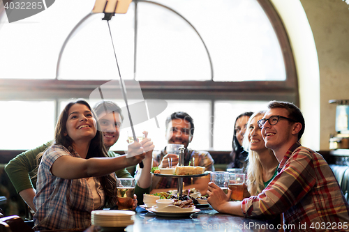 Image of happy friends with selfie stick at bar or pub