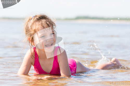 Image of Five-year girl lies in water in the shallows of the river, laughing and looking to the left