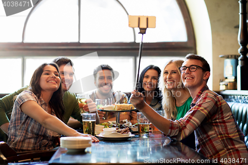Image of happy friends with selfie stick at bar or pub