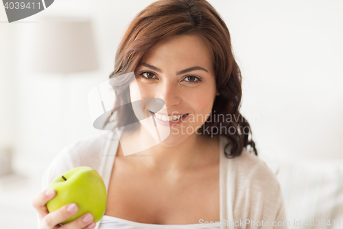 Image of smiling young woman eating green apple at home