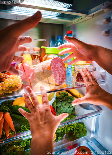 Image of Human hands reaching for food at night in the open refrigerator