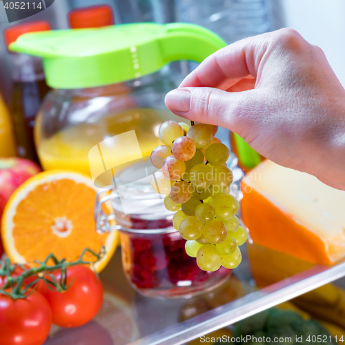 Image of Woman takes the bunch of grapes from the open refrigerator