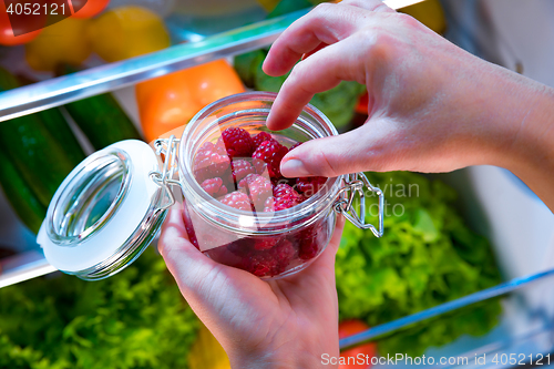 Image of Woman takes the fresh raspberries from the open refrigerator