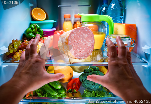 Image of Human hands reaching for food at night in the open refrigerator