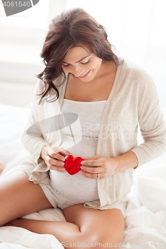 Image of happy pregnant woman with red heart in bed at home