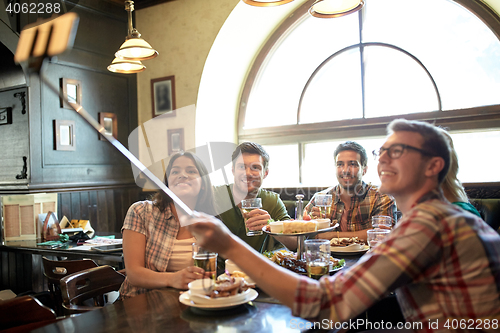 Image of happy friends with selfie stick at bar or pub