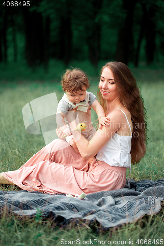 Image of Young beautiful mother sitting with her little son against green grass. Happy woman with her baby boy on a summer sunny day. Family walking on the meadow.