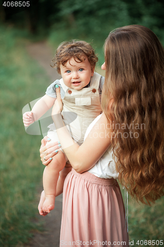 Image of Young beautiful mother hugging her little toddler son against green grass. Happy woman with her baby boy on a summer sunny day. Family walking on the meadow.