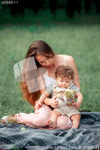 Image of Young beautiful mother sitting with her little son against green grass. Happy woman with her baby boy on a summer sunny day. Family walking on the meadow.