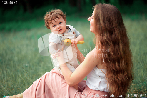 Image of Young beautiful mother sitting with her little son against green grass. Happy woman with her baby boy on a summer sunny day. Family walking on the meadow.