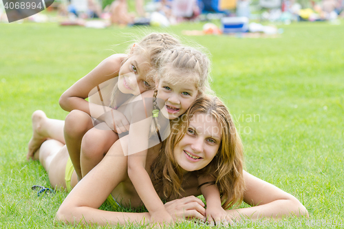 Image of Two daughters happily sit on mom back, hot summer day relaxing on the green lawn