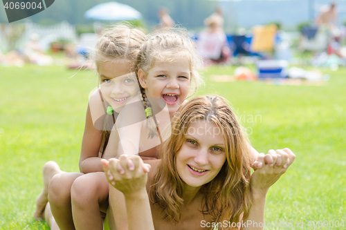 Image of Two daughters are sitting on the mother\'s back, hot summer day relaxing on the green lawn beautiful beaches