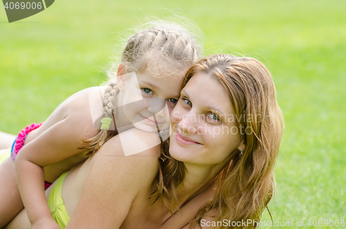 Image of Daughter lying on mother\'s back happily hugs her, against the background of green grass