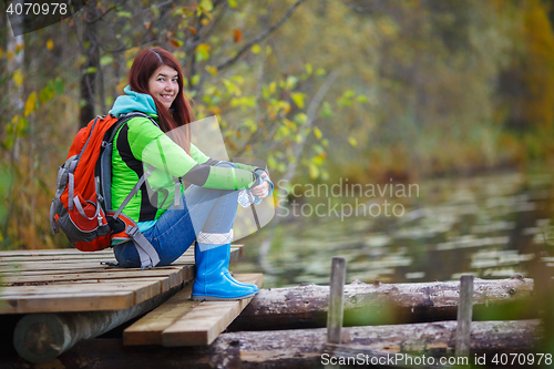 Image of Young brunette with backpack sitting on bridge by pond