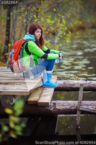 Image of Young brunette with travel backpack sits on bridge by pond