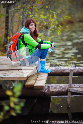 Image of Smiling brunette with travel backpack sits by pond in woods