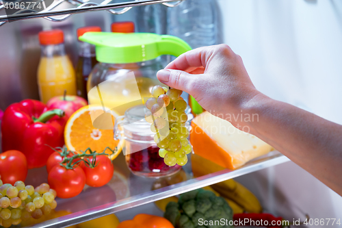 Image of Woman takes the bunch of grapes from the open refrigerator