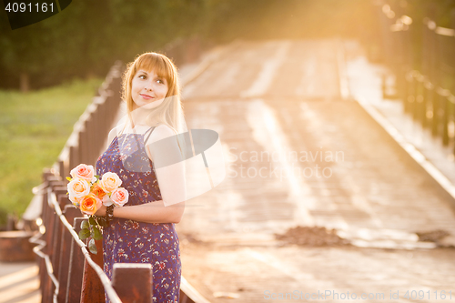 Image of The charming young girl standing on a rustic bridge at dawn sun with a bouquet of roses