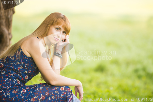 Image of Beautiful young girl sitting against a tree on the background of blurred green grass and looks into the distance