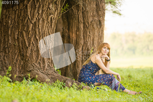 Image of Beautiful young woman sat on the roots of the old large tree and looked up dreamily