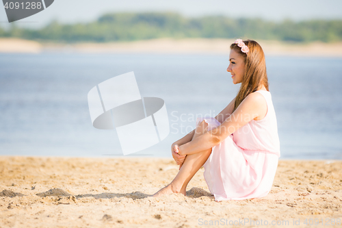 Image of Young girl in the summer light white dress sitting on the sandy beach by the river