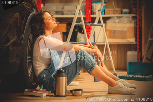 Image of auto mechanic woman in blue overalls resting near the wheel of t