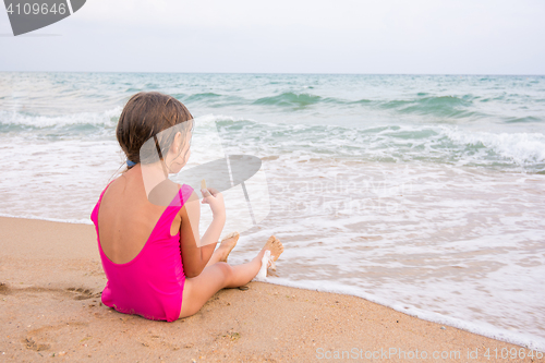 Image of The girl in the pink bathing suit sitting on the beach, eating a wafer and looks into the distance