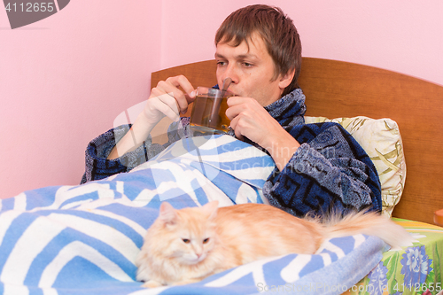 Image of A sick man lying in bed and drinking tea, lies next to the cat