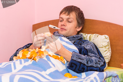 Image of A sick kid with a thermometer in her mouth lying in bed and eating tangerines