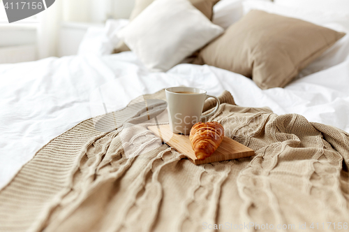 Image of coffee cup and croissant on plaid in bed at home