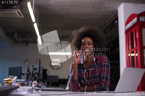 Image of black woman in modern office speeking on phone over earphones